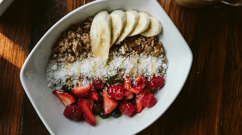 A white bowl of granola, sliced bananas, shredded coconut, strawberries, and raspberries on a wooden table.