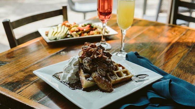 A waffle with whipped cream, caramelized bananas, and powdered sugar, with two mimosas and a salad.