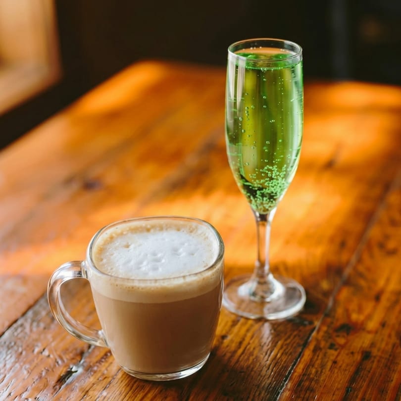 A frothy latte in a clear mug next to a champagne flute with a bubbling green drink.