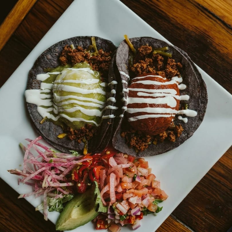 Two dark tacos with meat, guacamole, and sour cream, served with pico de gallo and avocado.