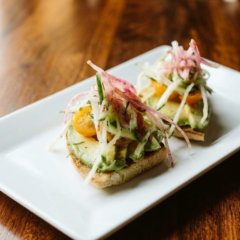 Two avocado toasts topped with cherry tomatoes, cucumber, and pickled red onions on a white plate.