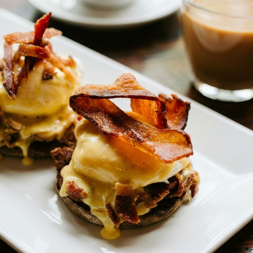 Two eggs Benedict with hollandaise sauce and crispy bacon on a white plate, with a coffee in the background.